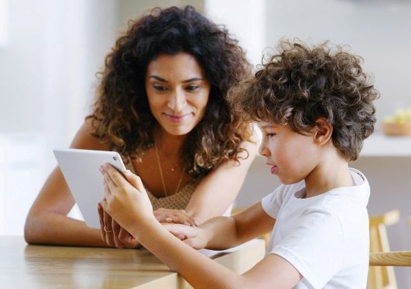 An adult woman watches as a young boy uses a tablet.