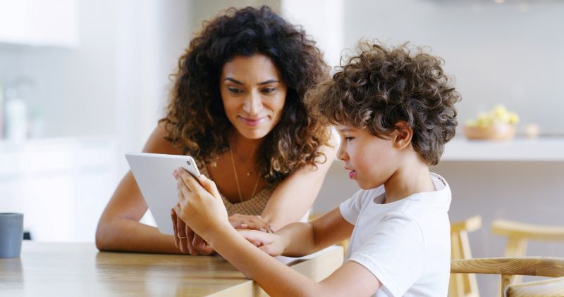 An adult woman watches as a young boy uses a tablet.