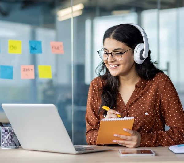A woman wearing headphones writes on a notepad while watching something on her laptop.