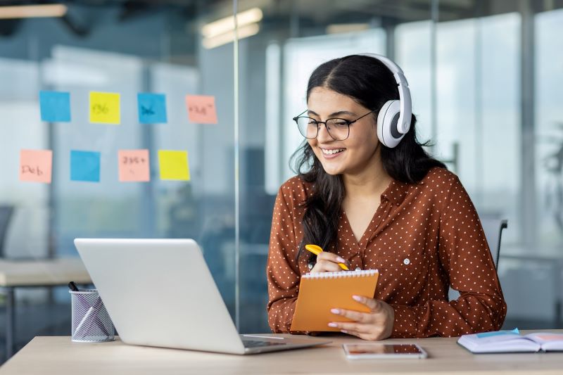 A woman wearing headphones writes on a notepad while watching something on her laptop.