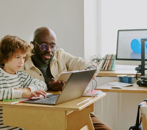 A teacher helps a student using a laptop in a classroom.