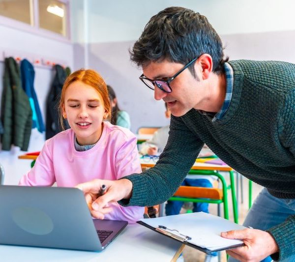 Teacher leaning over to assist a student working on a laptop in a bright classroom setting.