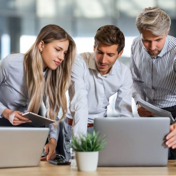 Three colleagues collaborating over laptops and tablets in a modern office setting with natural light.