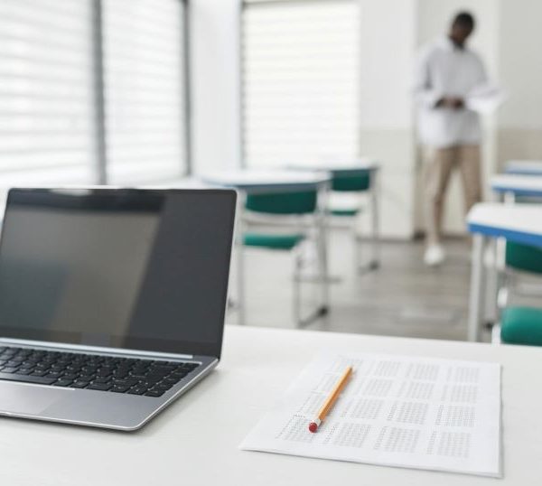 A desk with a laptop and a test in a classroom, with a teacher in the background a few rows back.