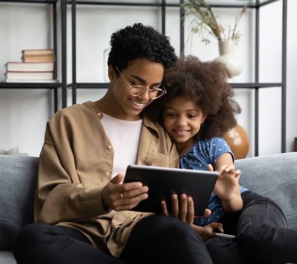 Smiling mother and daughter sitting on a couch together, enjoying a tablet screen in a cozy living room.