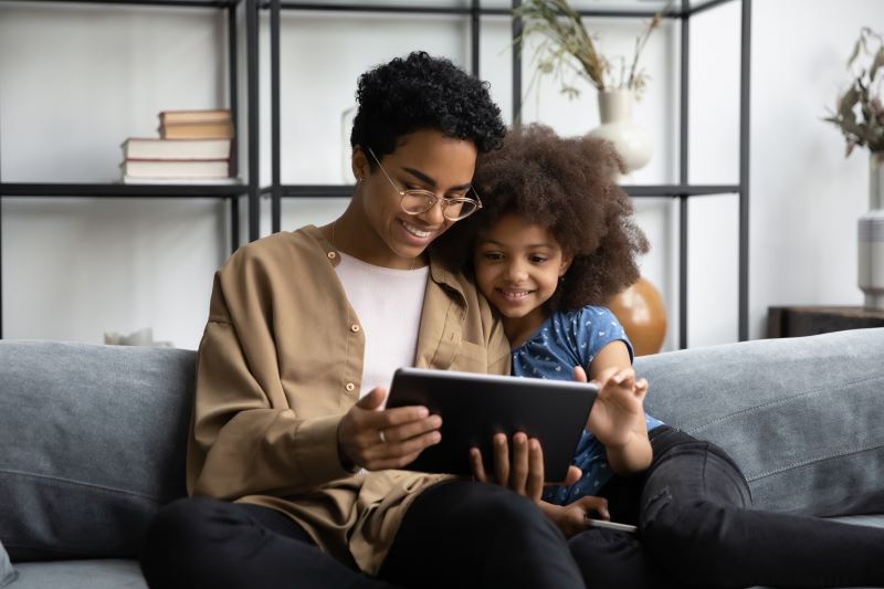 Smiling mother and daughter sitting on a couch together, enjoying a tablet screen in a cozy living room.