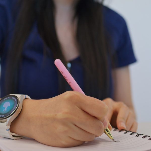 Person wearing a smartwatch writing notes with a pink pen in a spiral notebook on a wooden table.